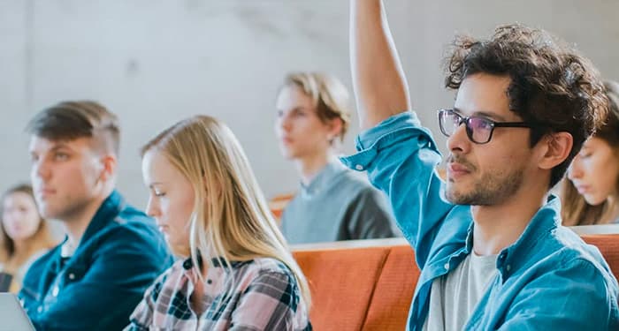 Male students in a classroom setting with others raising his hand to ask a question